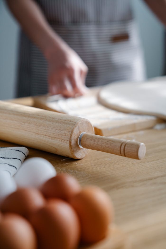 gallery-1 Hands preparing dough with a rolling pin and eggs on a wooden surface in a kitchen setting.