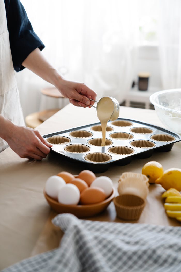 hero-img-02 A baker pours cake batter into a muffin tray with eggs and lemons on a table.